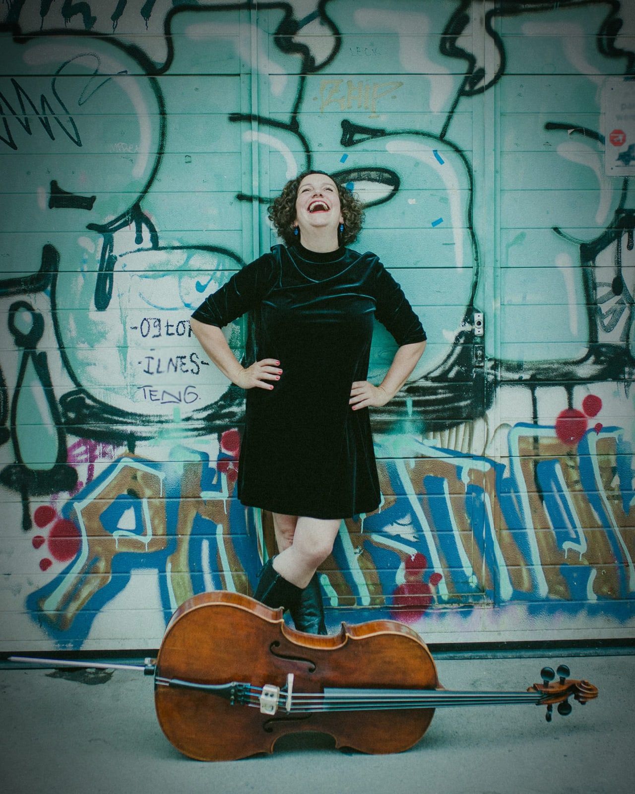 Claudia Herold lacht vor einer Graffitiwand, das Cello liegt vor ihr auf dem Boden